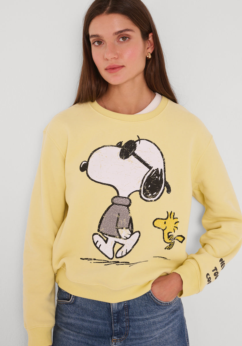 Eine junge Frau mit langen braunen Haaren trägt ein hellgelbes Princess Goes Hollywood Snoopy Sweatshirt aus Baumwolle, auf dem Snoopy mit Sonnenbrille und Pullover neben Woodstock läuft. Sie steht ruhig vor einem hellen Hintergrund, eine Hand in ihrer Jeanstasche.