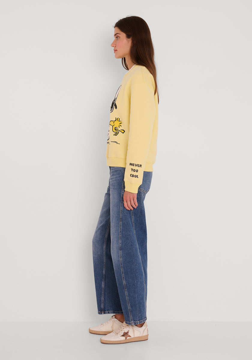 Eine Frau im Profil vor einer weißen Wand trägt ein gelbes grafisches "NEVER TOO COOL"-Sweatshirt von Princess Goes Hollywood, weit geschnittene Bluejeans und beigefarbene Turnschuhe. Ihr braunes Haar ist offen und ihre Arme sind an den Seiten verschränkt.