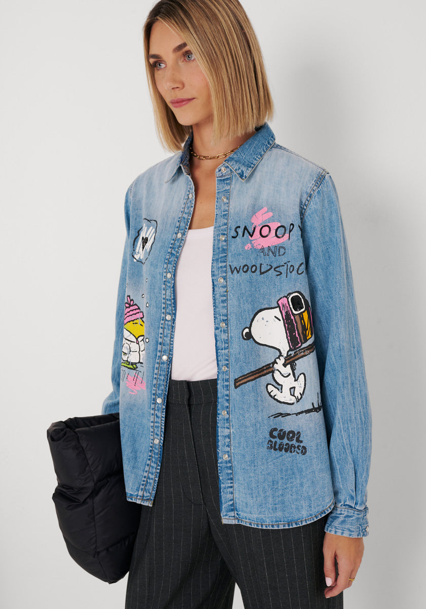 Eine Frau mit schulterlangem blondem Haar trägt ein blaues Baumwoll-Jeanshemd mit Snoopy- und Woodstock-Motiven, ein weißes Oberteil und eine dunkel gestreifte Hose. Sie hält eine schwarze Puffertasche in der Hand und steht vor einem weißen Hintergrund, wobei sie leicht zur Seite blickt.