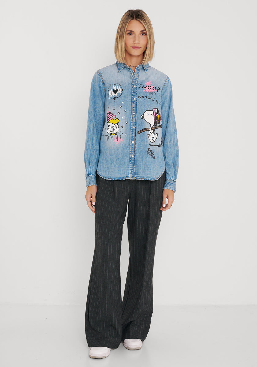 Eine Frau steht vor einer weißen Wand und trägt ein blaues Jeanshemd aus Baumwolle mit Snoopy-Aufdruck und Woodstock-Motiven, eine lockere schwarze Nadelstreifenhose und weiße Schuhe. Sie hat einen glatten blonden Bob und einen neutralen Gesichtsausdruck, die Arme hängen entspannt an den Seiten herab.