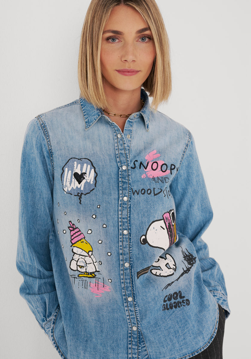 Eine Frau mit glattem blondem Haar trägt ein hellblaues Jeanshemd aus Baumwolle mit bunten Snoopy-Motiven, rosa Akzenten und den Aufschriften „SNOOP AND WOOD“ sowie „COOL SUMMER“. Sie blickt mit einem leichten Lächeln nach vorne und steht vor einem schlichten weißen Hintergrund.