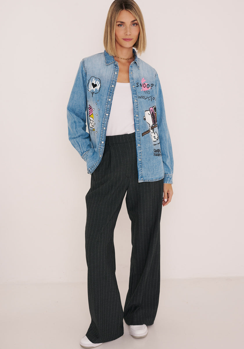 Eine Frau mit glattem blondem Haar trägt ein Princess Goes Hollywood-Jeanshemd mit Snoopy-Aufdruck über einem weißen Oberteil, gepaart mit einer weiten schwarzen Nadelstreifenhose und weißen Sneakers. Sie steht vor einem schlichten weißen Hintergrund und hat eine Hand in der Tasche.