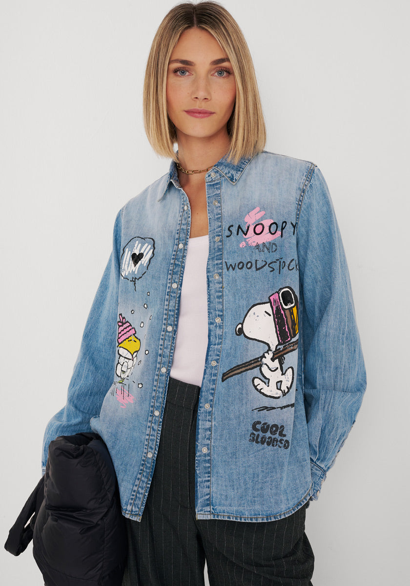Eine Frau mit Bob-Frisur trägt ein helles Jeanshemd aus Baumwolle mit Snoopy-Aufdruck und verspieltem Schriftzug. Darunter trägt sie ein weißes Trägertop und eine dunkle Nadelstreifenhose; in der Hand hält sie eine schwarze Jacke. Sie steht vor einem schlichten weißen Hintergrund und blickt in die Kamera.