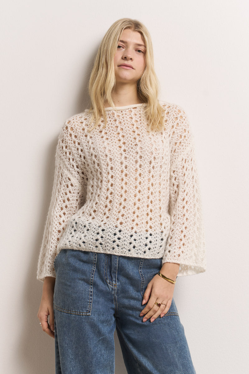 Eine junge Frau mit langen blonden Haaren trägt einen weißen, lockeren Ajourstrick-Pullover von Princess Goes Hollywood und eine hochgekrempelte Bluejeans. Sie steht an einer schlichten Wand, die Hand in der Tasche, und strahlt Selbstbewusstsein aus; ein einfaches goldenes Armband vervollständigt ihren stilvollen Look.