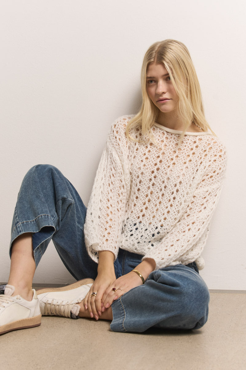 Eine junge Frau mit langen blonden Haaren sitzt an einer weißen Wand auf einem hellbraunen Boden. Sie trägt lässige Bluejeans, weiße Turnschuhe und einen luftigen Ajourstrick-Pullover von Princess Goes Hollywood. Sie schaut ruhig in die Kamera, hat ein Knie hoch und die Arme auf den Beinen.