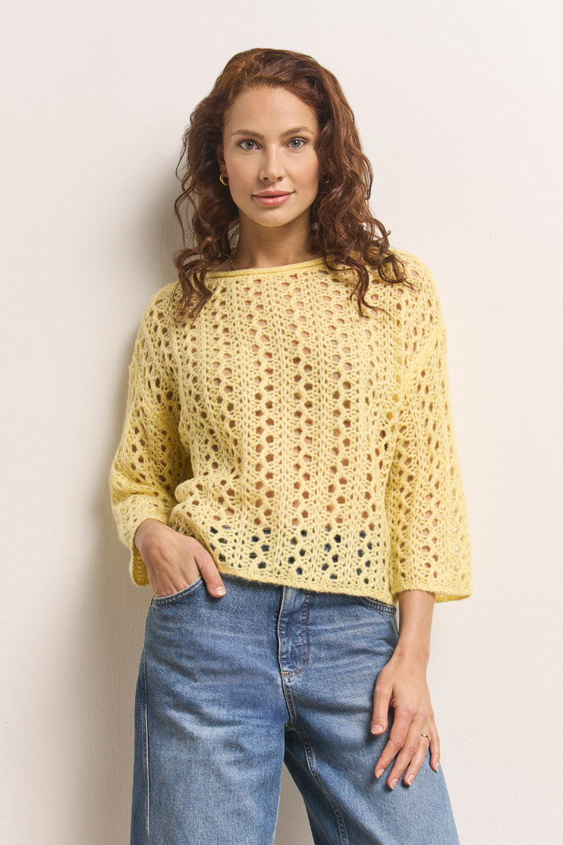 Eine Frau mit gewellten roten Haaren trägt einen gelben Princess Goes Hollywood Ajourstrick Pullover Damenpullover in Merino-Alpaka-Mix und blaue Jeans und steht vor einer cremefarbenen Wand. Sie schaut mit einem leichten Lächeln in die Kamera, eine Hand in der Tasche.