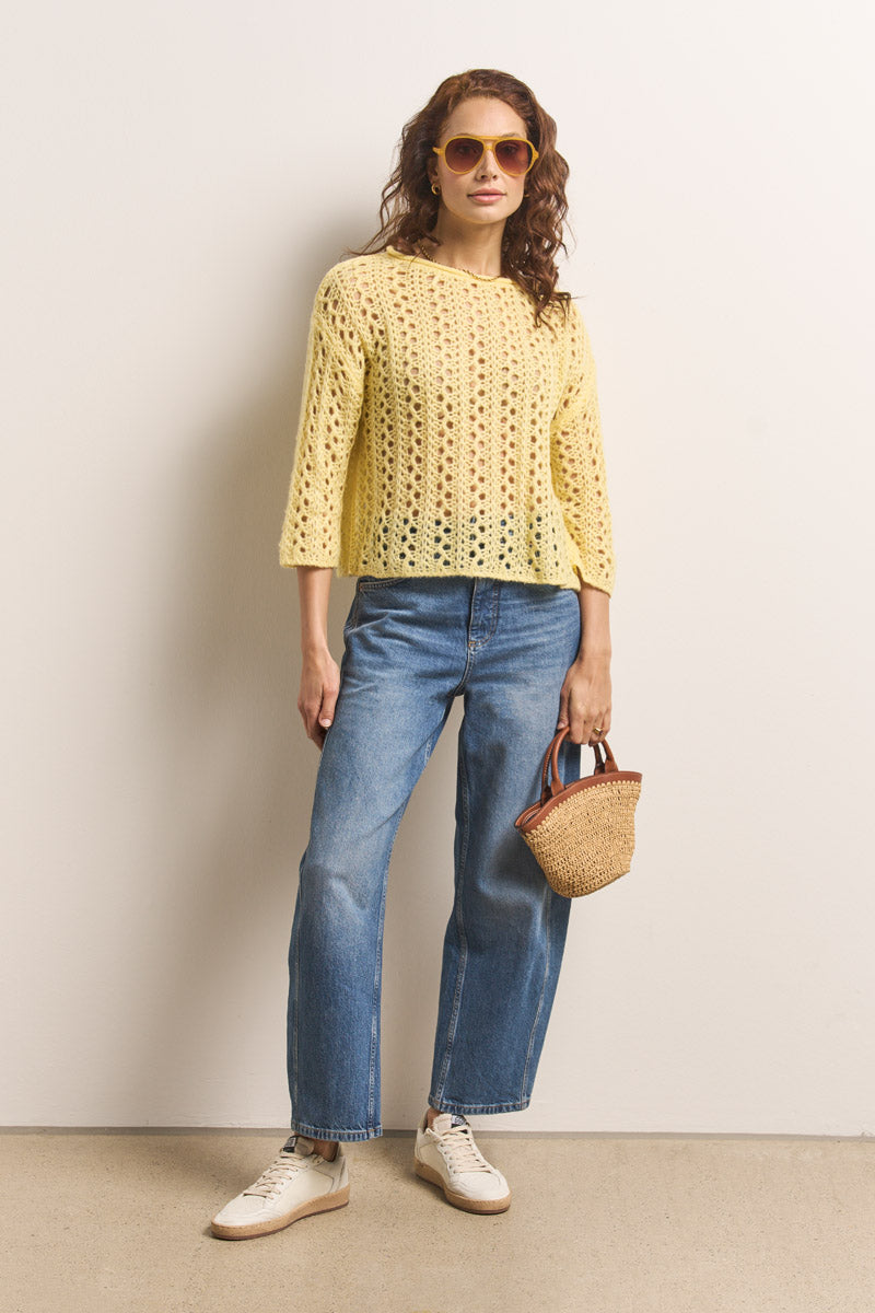 Eine Person mit gewelltem, schulterlangem braunem Haar steht vor einer schlichten Wand und trägt einen gelben Häkelpulli von Okkia, lässige Bluejeans, weiße Turnschuhe und eine große gelbe Okkia Aviator Sonnenbrille mit UV 400 Schutz. In der Hand hält sie eine geflochtene Strohhandtasche.