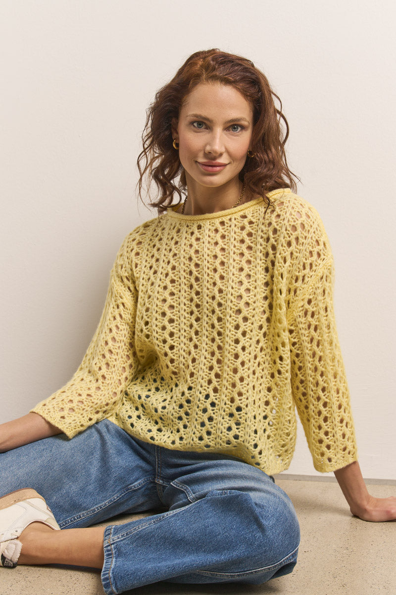 Eine Frau mit gewelltem rotem Haar lächelt sanft gegen eine weiße Wand. Sie trägt einen hellgelben Princess Goes Hollywood Ajourstrick-Pullover aus weichem Merino-Alpaka-Mix, Dreiviertelärmel und blaue Jeans. Ein Knie ist angewinkelt, die Hände ruhen auf ihren Beinen.