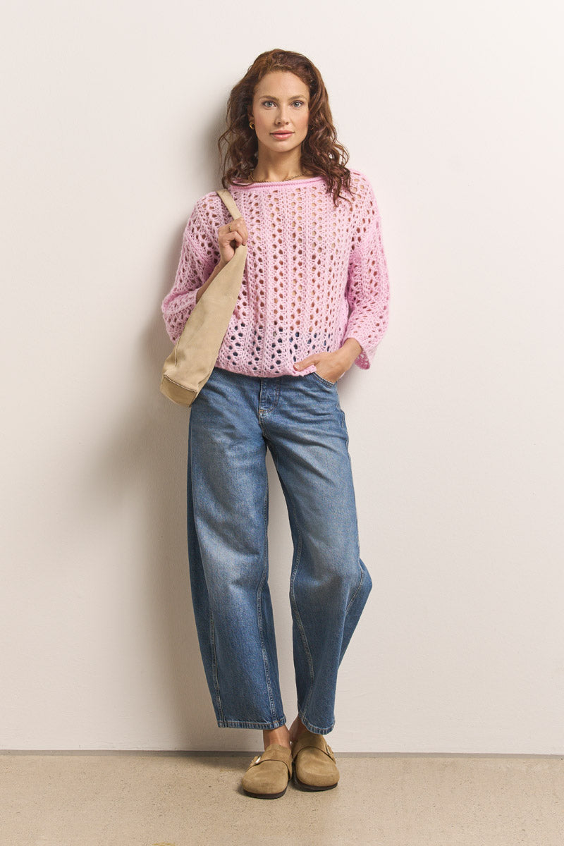 Eine Frau mit gewelltem braunem Haar steht vor einer weißen Wand. Sie trägt einen hellrosa Princess Goes Hollywood Damen Pullover im Ajourstrick aus Merino-Alpaka-Mix, blaue Jeans mit weitem Bein, hellbraune Slipper und eine beige Umhängetasche. Sie schaut selbstbewusst in die Kamera.