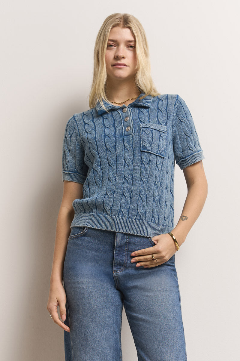 Eine junge Frau mit glattem blondem Haar trägt einen blauen, kurzärmeligen Princess Goes Hollywood Zopfstrick-Pullover aus Baumwolle mit Brusttasche und stone-wash Bluejeans und steht mit einem entspannten, leichten Lächeln vor einer cremefarbenen Wand.