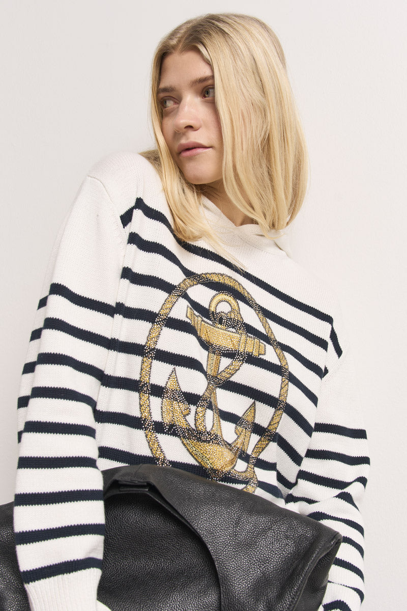 Eine Frau mit glattem blondem Haar trägt einen weißen, gestreiften Kapuzenpullover aus 100% Baumwolle mit einem Ankermotiv von Princess Goes Hollywood. Sie blickt zur Seite und hält ein schwarzes, strukturiertes Accessoire in den Händen.