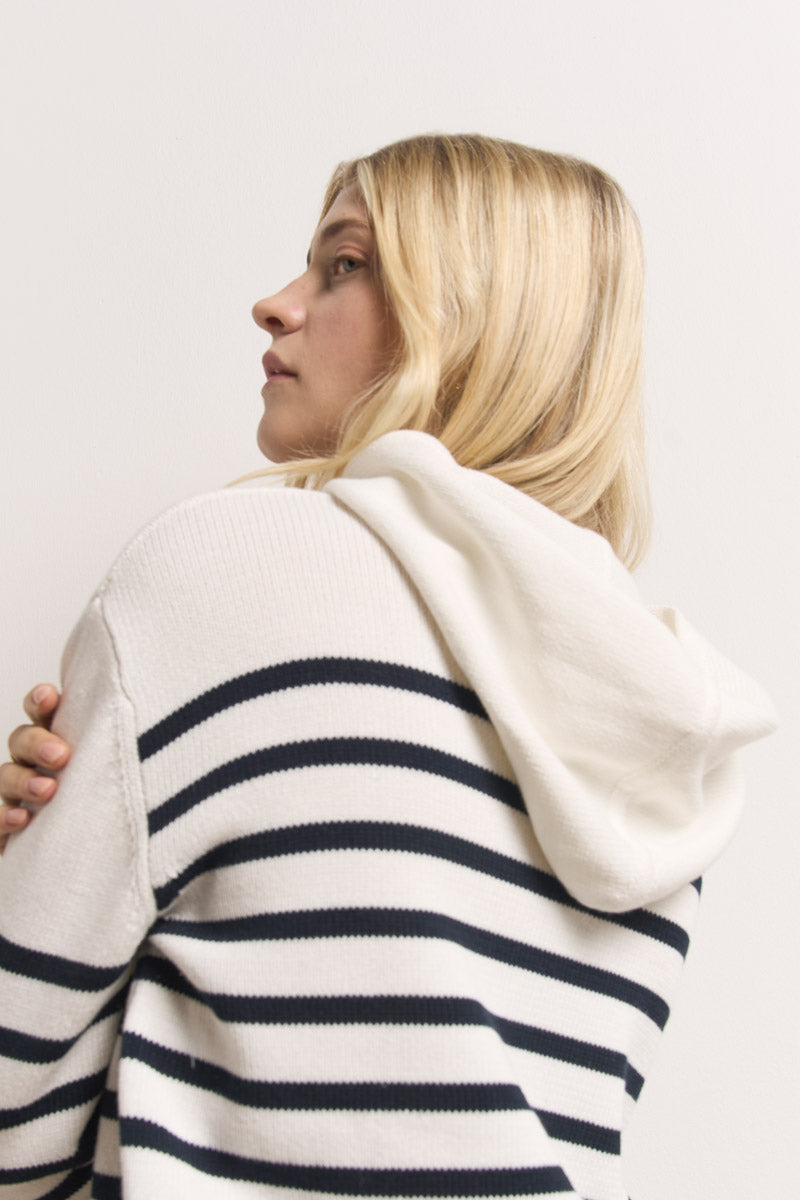 Eine Person mit hellblondem Haar trägt einen Princess Goes Hollywood gestrickter Hoodie aus 100 % Baumwolle mit schwarzen Querstreifen und einer großen weißen Kapuze und blickt von der Kamera weg auf einen schlichten, hellen Hintergrund.