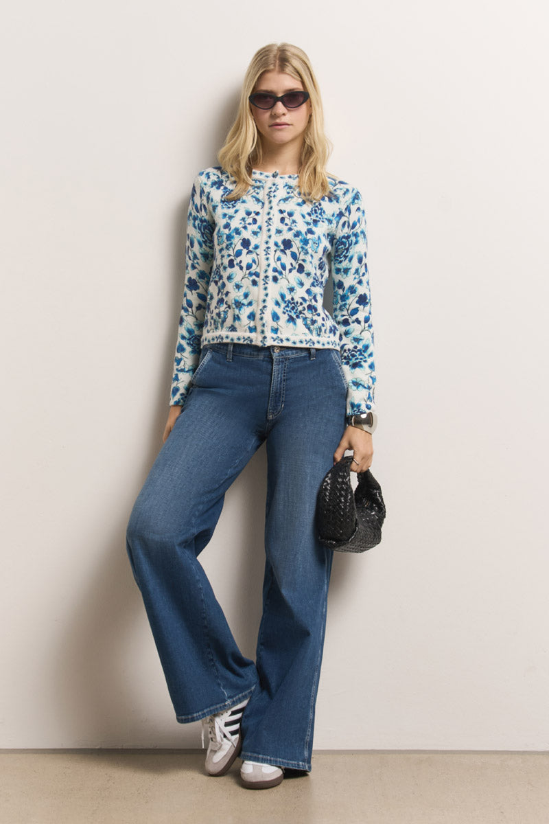 Eine Frau mit Sonnenbrille posiert vor einer schlichten Wand und trägt eine blaue Princess Goes Hollywood Blumen-Print Strickjacke, Jeans mit weitem Bein und weiße Turnschuhe. Sie trägt eine kleine schwarze, strukturierte Handtasche und ihr glattes blondes Haar vervollständigt den entspannten Look.