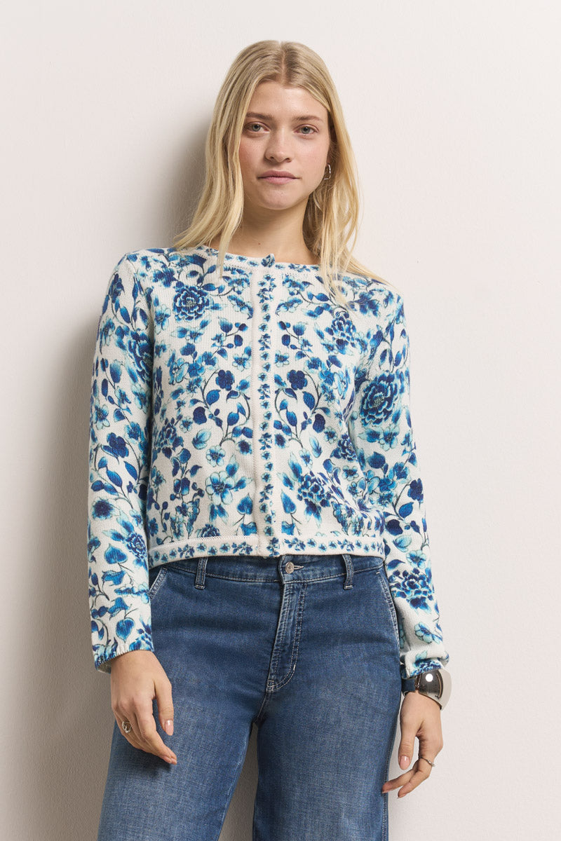 Eine Frau mit langen blonden Haaren trägt eine weiße Baumwoll-Strickjacke mit blauem Blumen-Print von Princess Goes Hollywood. Dazu kombiniert sie hoch taillierte blaue Jeans und steht vor cremefarbenem Hintergrund.