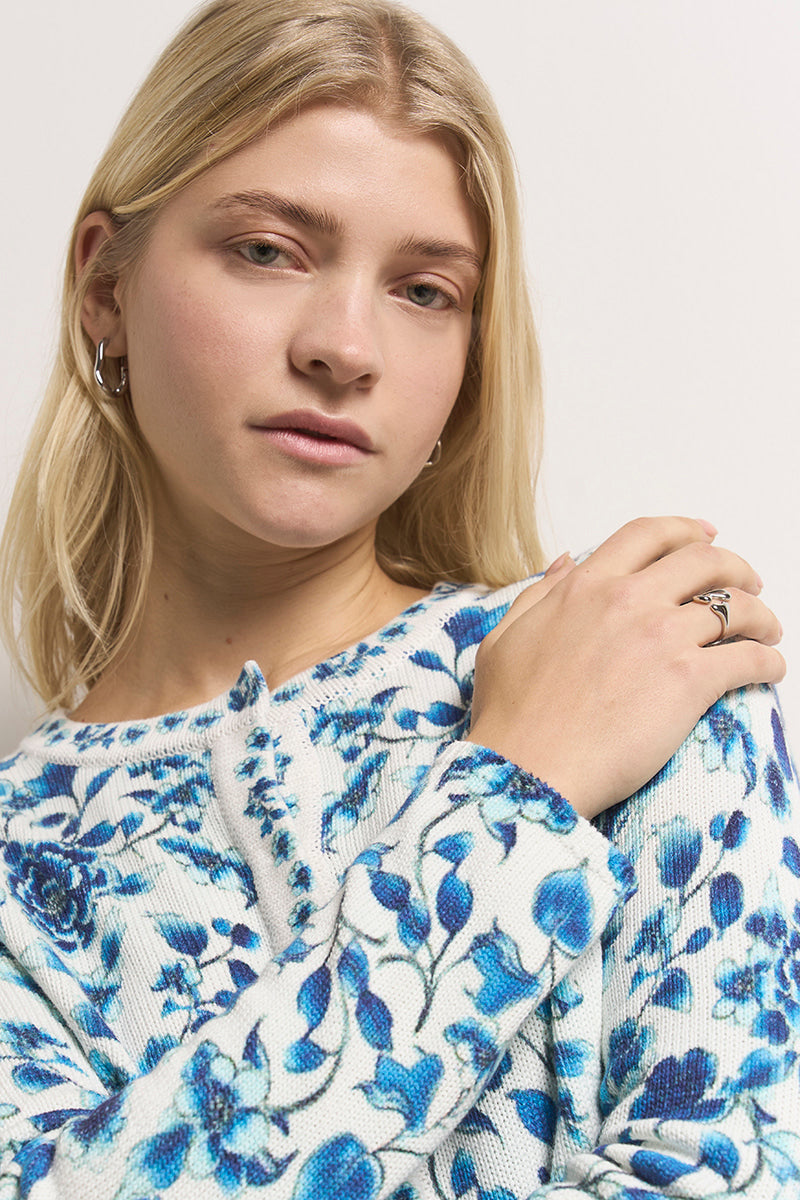 Eine junge Frau mit glattem blondem Haar trägt Ohrringe und eine weiße Princess Goes Hollywood Baumwolle Strickjacke mit blauem Blumendruck. Sie posiert ruhig mit einer Hand auf ihrer Schulter, die silberne Ringe zeigt, vor einem schlichten weißen Hintergrund.