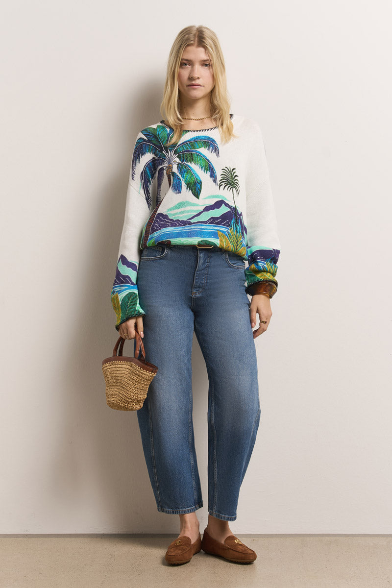 Eine Frau mit glattem blondem Haar und neutralem Gesichtsausdruck trägt einen weißen Princess Goes Hollywood Pullover Baumwolle mit buntem Karibik-Print, blaue High-Waisted-Jeans, braune Loafers und trägt eine kleine Strohhandtasche vor einer hellen Wand.