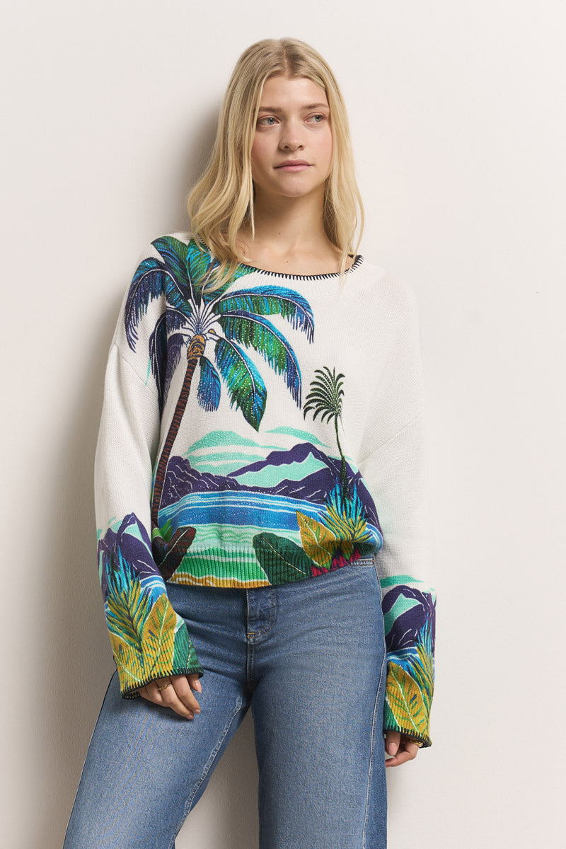 Eine junge Frau mit langen blonden Haaren trägt einen Princess Goes Hollywood-Baumwollpullover mit einem farbenfrohen karibischen Aufdruck von Palmen, Bergen und Meereswellen. Sie steht vor einem hellen Hintergrund und schaut entspannt zur Seite, eine Hand in ihrer Jeans.