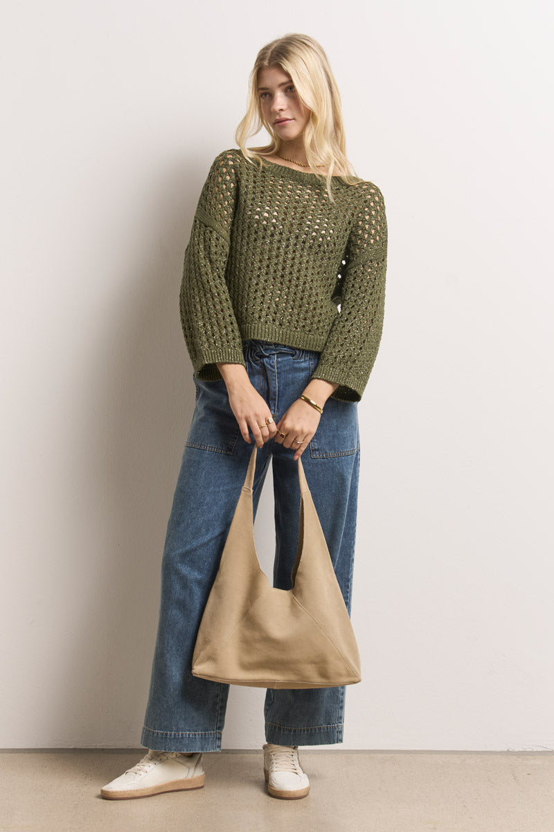 Eine Frau steht an einer schlichten Wand. Sie trägt einen lockeren olivgrünen Lochstrickpullover aus Bio-Baumwolle von Princess Goes Hollywood, eine weit geschnittene Bluejeans und weiße Turnschuhe. Sie trägt eine hellbraune Schlabberhandtasche und hat lockeres, seitlich gescheiteltes blondes Haar.