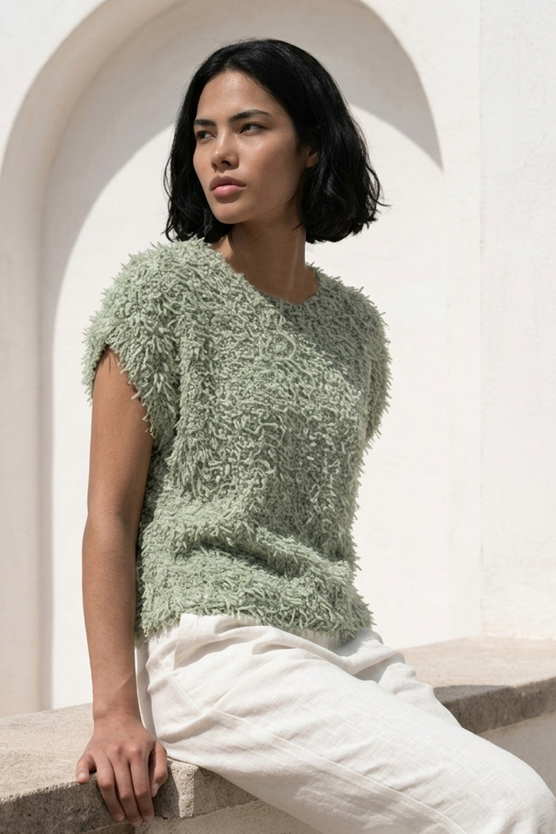 Eine Frau mit kinnlangem schwarzem Haar trägt einen blassgrünen Princess Goes Hollywood ärmellos Pullunder aus Effektgarn und eine weite weiße Hose. Sie sitzt auf einem Steinsims unter sonnenbeschienenen Bögen und blickt mit ruhigem Blick nach links.