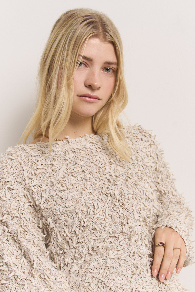 Eine junge Frau mit langen blonden Haaren und heller Haut steht vor einer weißen Wand, trägt einen Princess Goes Hollywood Effektgarn Pullover mit Rundhalsausschnitt und einen goldenen Ring und blickt ruhig zur Seite.