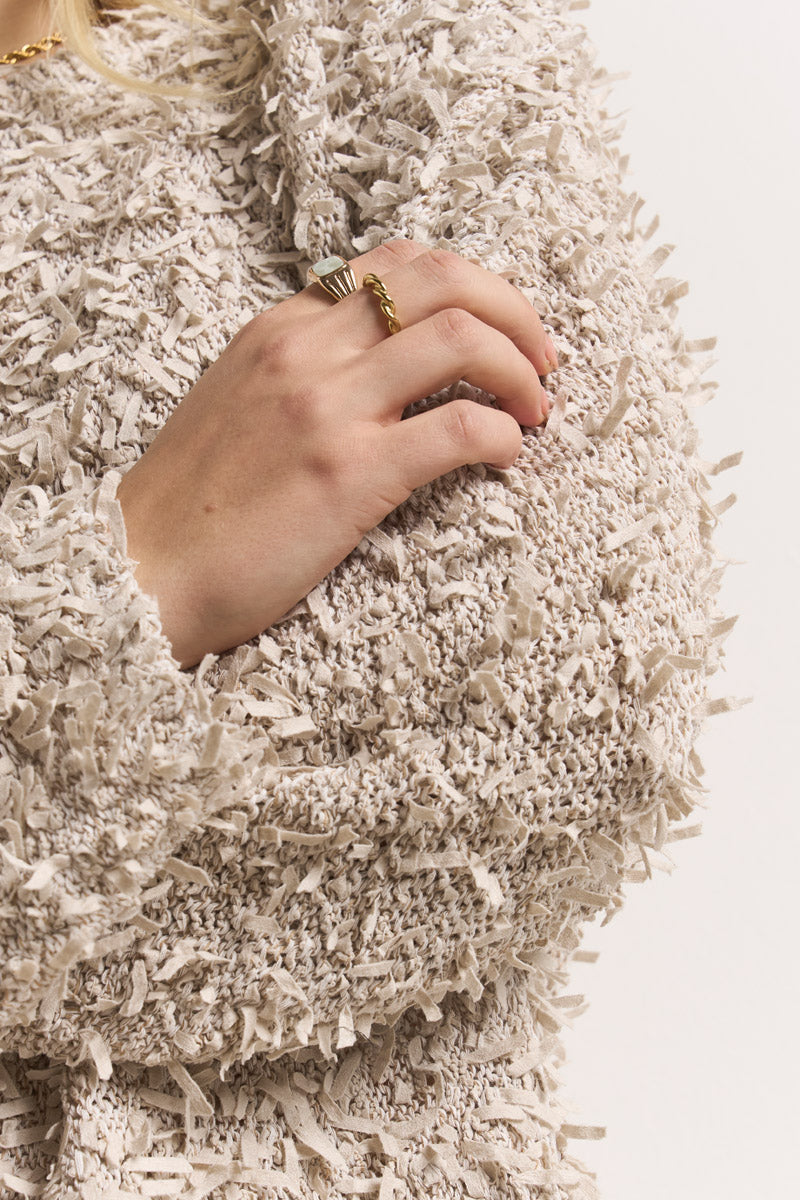 Nahaufnahme einer Hand, die auf einem strukturierten, cremefarbenen Princess Goes Hollywood Effektgarn-Pullover mit Fransendetails ruht; die Person trägt goldene und silberne Ringe und hat kurze, natürliche Nägel. Der einfarbige Hintergrund ist hell.