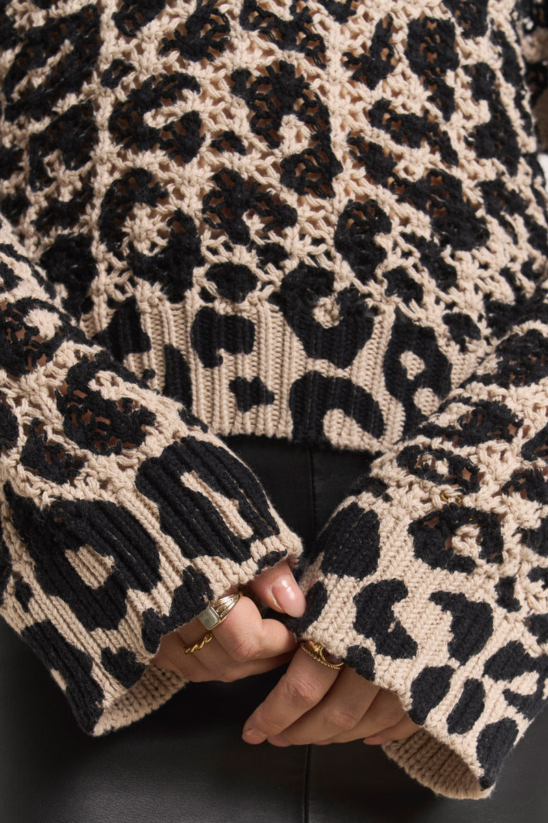 Eine Nahaufnahme zeigt den Oberkörper und die Hände einer Person, die einen beige-schwarzen Häkelpulli mit Leo-Print von Princess Goes Hollywood trägt. Die langen Ärmel verdecken einen Teil der gefalteten Hände, die mit goldenen Ringen verziert sind, gepaart mit einer schwarzen Hose.