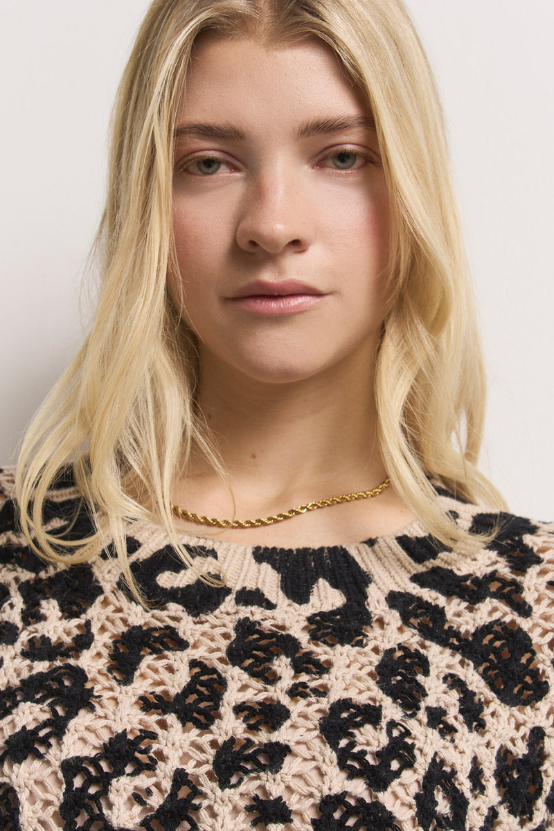 Eine Person mit langen blonden Haaren und heller Haut blickt in die Kamera. Sie trägt eine goldene, gedrehte Kettenhalskette und ein beigefarbenes Baumwolle-Top mit Leo-Print in Häkeloptik von Princess Goes Hollywood. Sie haben einen neutralen Ausdruck vor einem schlichten weißen Hintergrund.