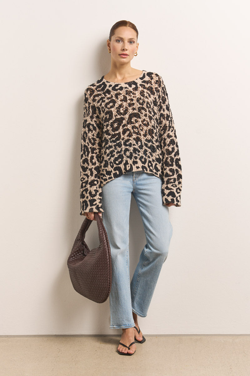 Eine Frau trägt einen lockeren, langärmeligen Leo-Print gehäkelter Pullover von Princess Goes Hollywood, hellblaue Jeans mit geradem Bein, schwarze Sandalen und eine strukturierte dunkelbraune Handtasche. Ihr Haar ist hochgesteckt und sie trägt kleine Ohrringe.