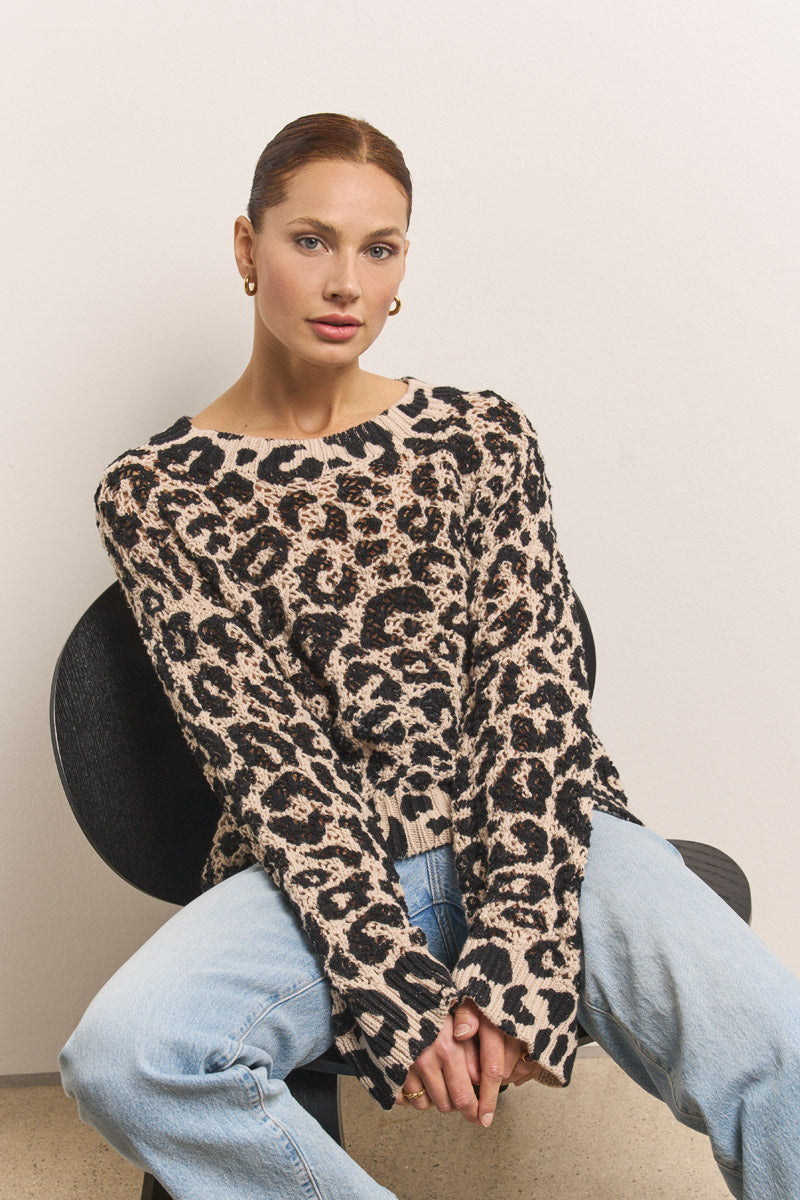 Eine Frau mit zurückgebundenem Haar und goldenen Ohrringen sitzt auf einem schwarzen Stuhl, trägt hellblaue Jeans und einen Princess Goes Hollywood 100% Baumwolle Leo-Print gehäkelter Pullover, mit einem entspannten, neutralen Gesichtsausdruck und gefalteten Händen im Schoß.