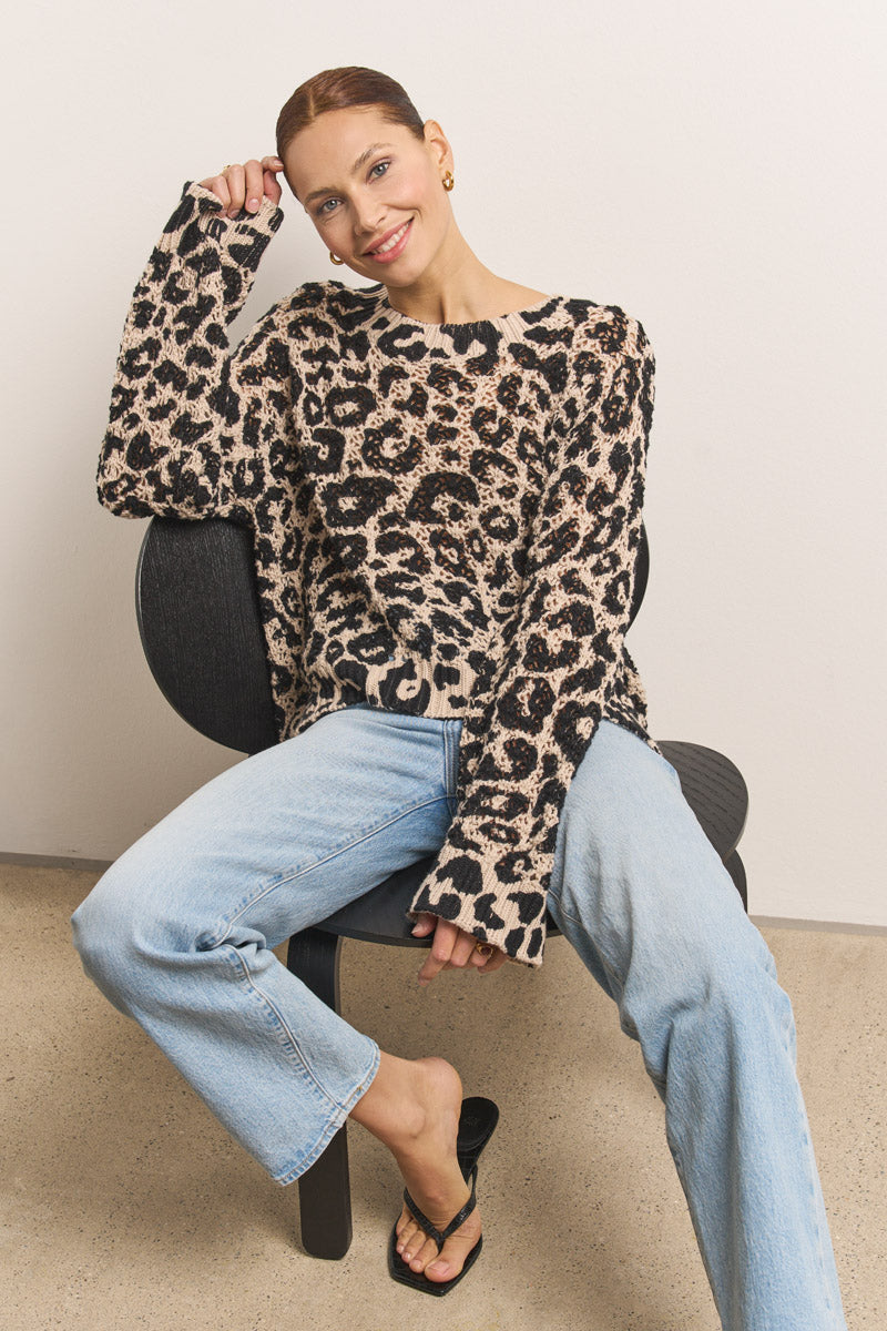 Eine lächelnde Frau mit zurückgekämmtem Haar sitzt auf einem schwarzen Stuhl und posiert lässig in einem Princess Goes Hollywood 100% Baumwolle Leo-Print gehäkelter Pullover, hellblauen Jeans und schwarzen Sandalen vor einem schlichten Hintergrund.