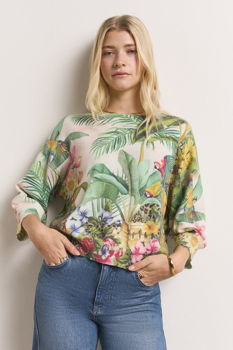 Eine Frau mit langen blonden Haaren trägt einen Princess Goes Hollywood-Pullover mit Dschungelaufdruck, grünen Blättern und bunten Blüten. Sie steht vor einer hellen Wand, eine Hand in der Jeanstasche und mit einem entspannten Gesichtsausdruck.