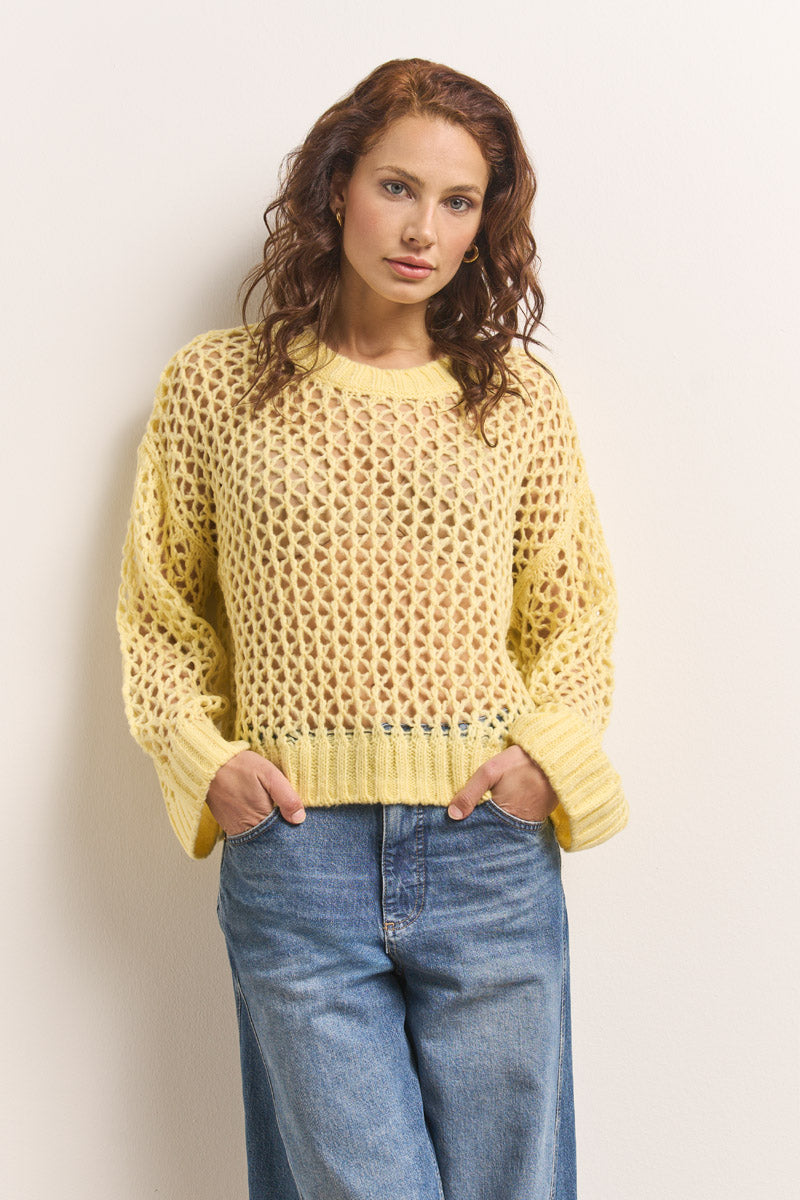 Eine Frau mit gewelltem, rötlich-braunem Haar posiert an einer weißen Wand. Sie trägt selbstbewusst einen lockeren gelben Princess Goes Hollywood Lochstrick-Pullover über einem hellen Oberteil und blaue, hochgekrempelte Jeans, die Hände in den Taschen.