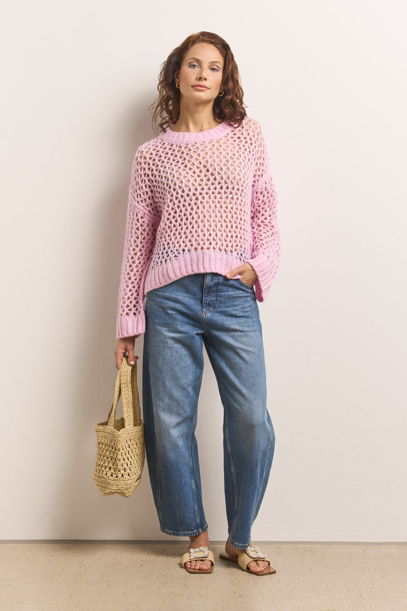 Eine Frau mit lockigem rotem Haar trägt einen lockeren, hellrosa "Princess Goes Hollywood"-Pullover über einem weißen Oberteil, blaue Jeans mit weitem Bein und beigefarbene Sandalen. Sie hält eine geflochtene Tragetasche aus Stroh in der Hand und steht vor einer schlichten, hellen Wand, die der Kamera zugewandt ist.