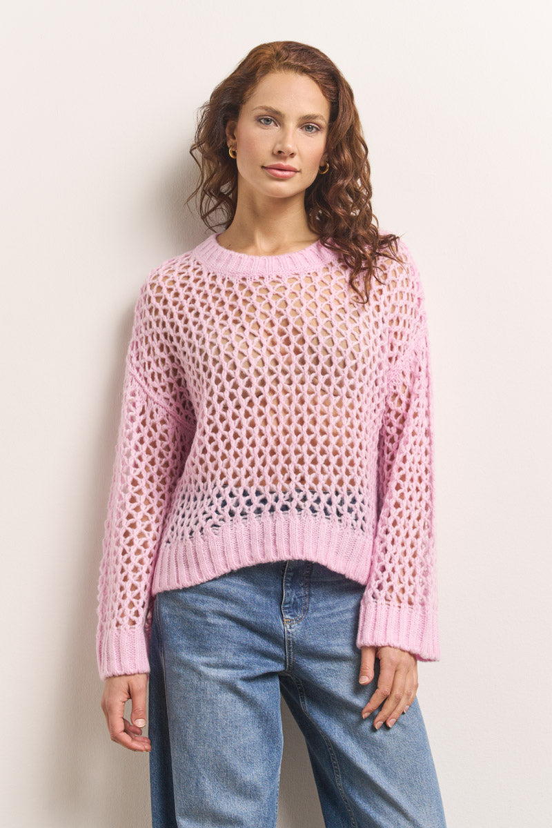 Eine Frau mit gewelltem braunem Haar trägt einen hellrosa Lochstrick Damen Pullover von Princess Goes Hollywood über einem nudefarbenen Oberteil und blauen Jeans. Sie steht vor einer schlichten Wand und blickt mit einer Hand an der Seite und einem entspannten Gesichtsausdruck nach vorne.