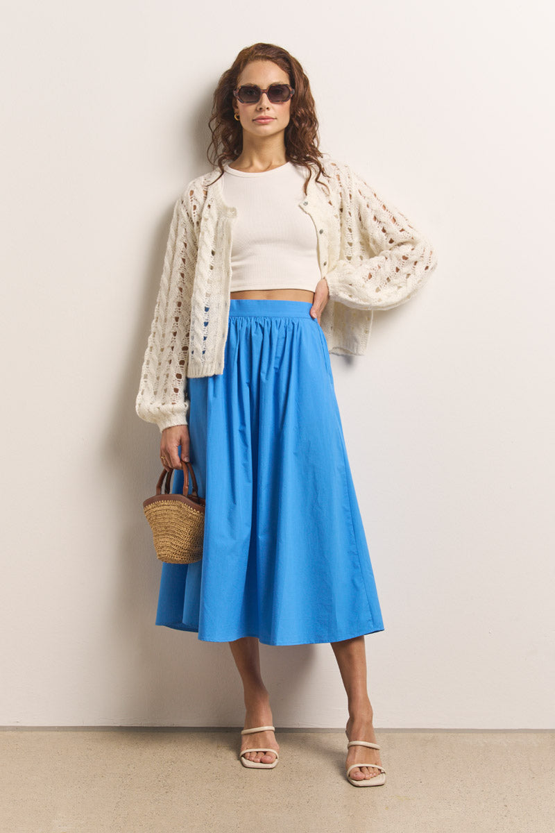 Eine Frau mit langen gewellten Haaren, die die cremefarbene Lochstrick-Strickjacke von Princess Goes Hollywood über einem weißen Crop-Top, einer Sonnenbrille, einem leuchtend blauen Midirock und weißen Sandalen trägt, hält eine geflochtene Strohhandtasche vor einer hellen Wand.