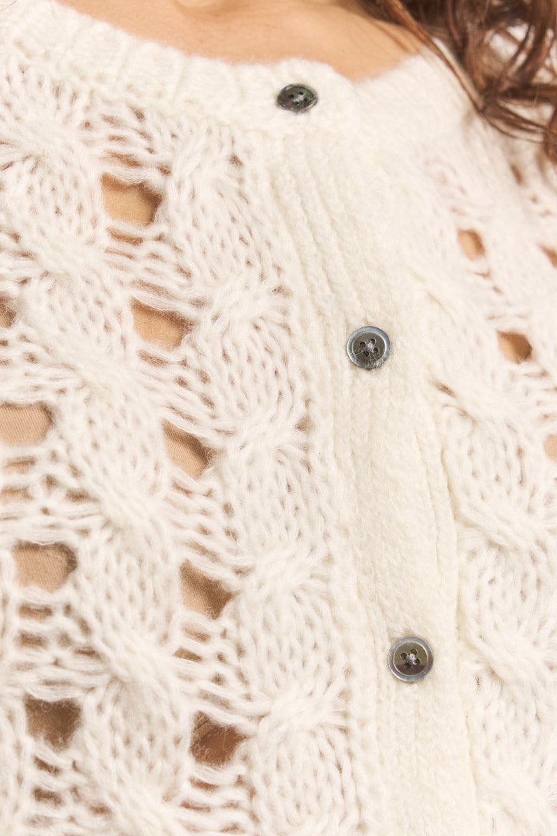 Nahaufnahme einer cremefarbenen Strickjacke von Princess Goes Hollywood in Lochstrick, gefertigt aus weichem Merino-Alpaka-Mix. Dunkle, runde Knöpfe säumen die gerippte Knopfleiste, durch das offene Gestrick ist Haut zu sehen.