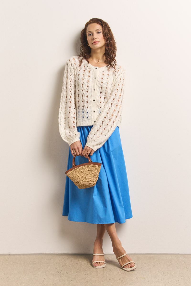 Eine Frau steht in einer cremefarbenen Princess Goes Hollywood Lochstrick-Strickjacke, einem hellblauen Midirock und hellbraunen Gleitsandalen vor einer weißen Wand und hält eine kleine geflochtene Strohhandtasche in der Hand. Ihr braunes gewelltes Haar fällt ihr über die Schultern, während sie nach oben blickt.