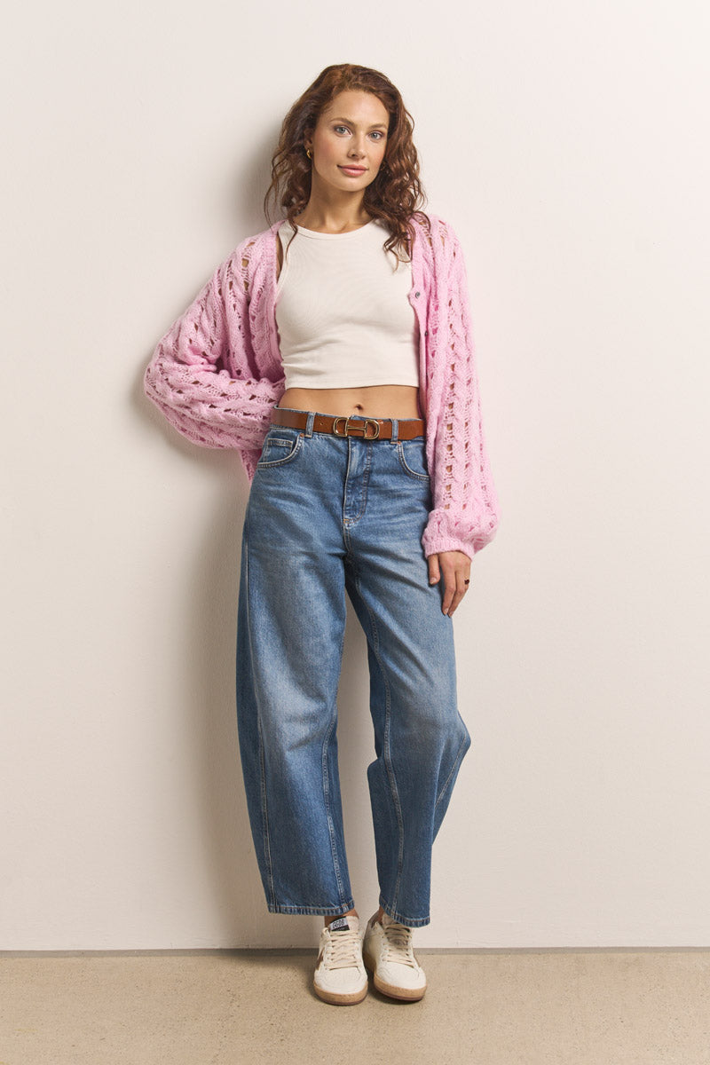 Eine Frau mit gewelltem braunem Haar steht vor einer hellen Wand. Sie trägt eine rosa Lochstrick-Strickjacke von Princess Goes Hollywood über einem weißen Crop-Top, blaue High-Waisted-Jeans mit braunem Gürtel und weiße Turnschuhe und strahlt entspanntes Selbstbewusstsein aus.