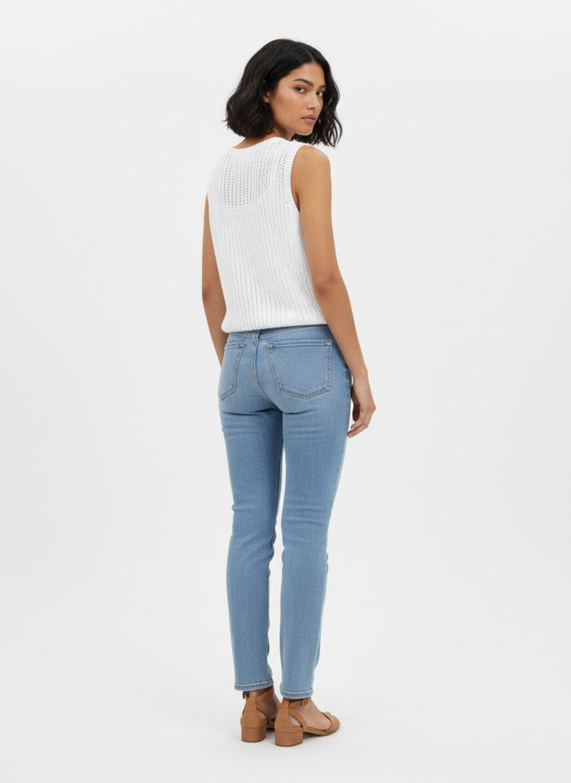 Eine Frau mit kinnlangen dunklen Haaren, die sich abwendet und über die Schulter schaut, trägt einen Princess Goes Hollywood weißen Lochstrick-Pullunder aus Baumwolle mit Rundhalsausschnitt, hellblaue Skinny Jeans und hellbraune flache Schuhe vor einem weißen Hintergrund.