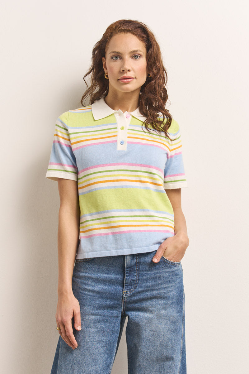 Eine Frau mit gewelltem rotem Haar trägt ein pastellfarben gestreiftes Baumwoll-Kaschmir-Poloshirt mit blauen Knöpfen von Princess Goes Hollywood und steht selbstbewusst mit einer Hand in der Tasche ihrer blauen Jeans vor einer cremefarbenen Wand.