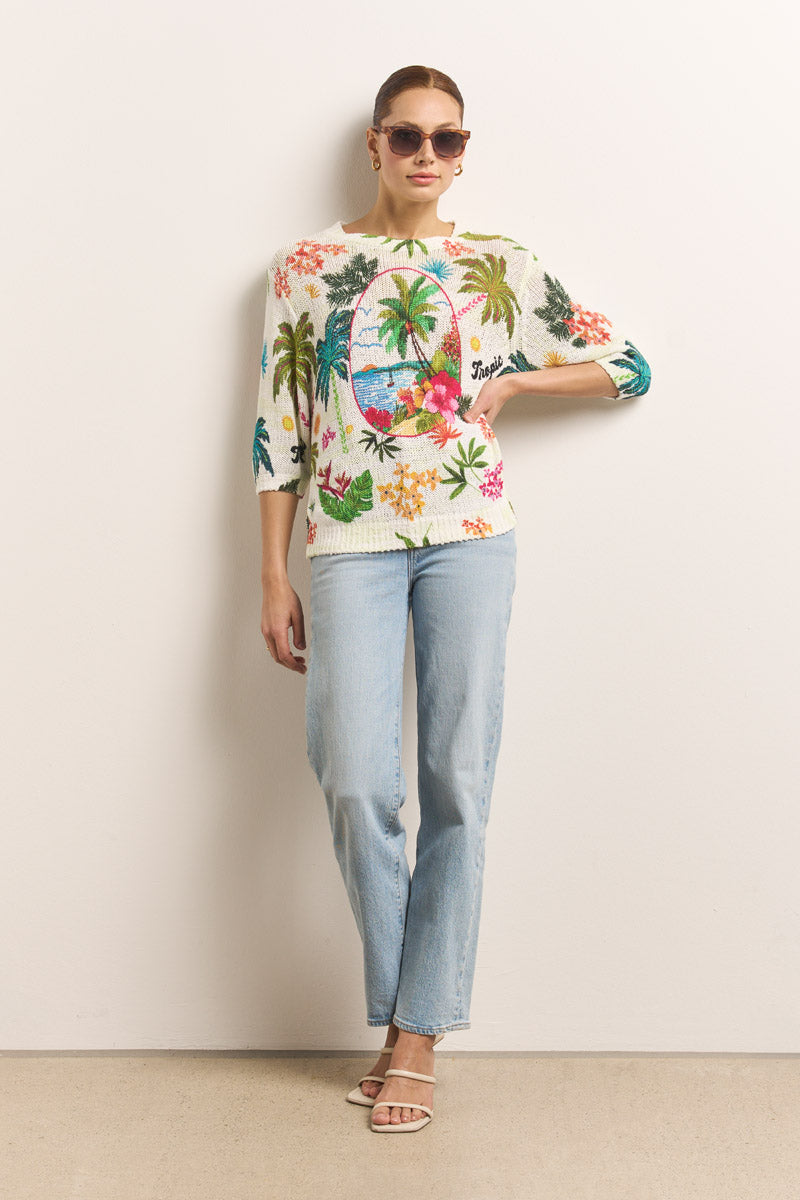 Eine Frau mit zurückgebundenem Haar, einen Arm in die Hüfte gestemmt, trägt eine Okkia Square-Frame Sonnenbrille mit UV 400-Schutz, einen Pullover mit tropischen Blumen und Palmen, hellblaue Jeans und weiße Sandalen mit Absätzen vor einer schlichten Wand.