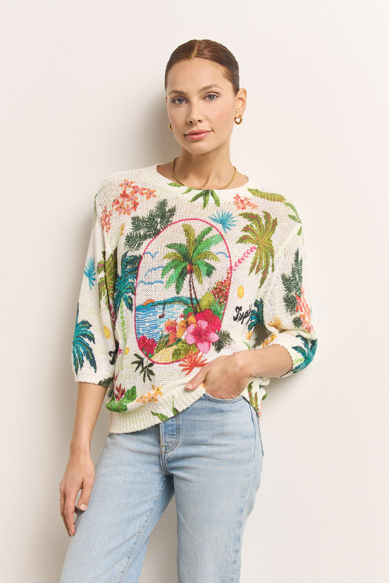 Eine Frau mit heller Haut und zurückgebundenen braunen Haaren steht vor einer weißen Wand. Sie trägt goldene Ohrringe, hellblaue Jeans und einen Princess Goes Hollywood Palmenprint Pullover aus Bio-Baumwolle mit Palmen, Blumen und einer Meeresszene.