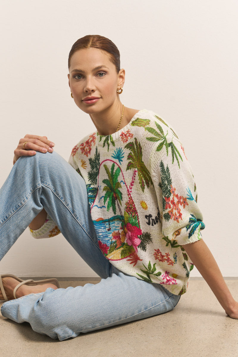 Eine Frau mit heller Haut und kastanienbraunem Haar in einem Dutt sitzt auf dem Boden vor einer weißen Wand. Sie trägt goldene Ohrringe, hellblaue Jeans, Sandalen und einen Princess Goes Hollywood Damen Pullover mit Palmenprint aus Bio-Baumwolle.