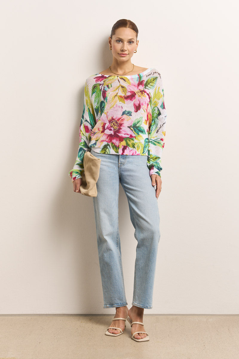 Eine Frau steht vor einer schlichten Wand und trägt einen Princess Goes Hollywood bunte Blumenprint Pullover und hellblaue Jeans mit geradem Bein. Sie hat eine beigefarbene Clutch in der Hand, trägt beigefarbene Sandalen, hat ihr Haar zurückgesteckt und schmückt sich mit goldenen Reifohrringen.
