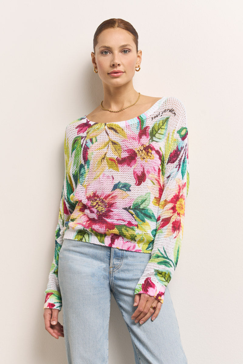 Eine Frau mit glattem braunem Haar trägt einen Princess Goes Hollywood Blumenprint-Pullover mit großen, lebhaften Blumenmustern, dazu hellblaue Jeans und goldene Ohrringe. Sie steht vor einer schlichten, hellen Wand und hat einen neutralen Gesichtsausdruck.