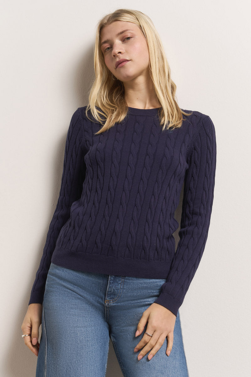 Eine Frau mit glattem blondem Haar trägt einen marineblauen Princess Goes Hollywood Zopfstrick-Pullover mit Rundhalsausschnitt und hellblaue Jeans. Sie steht vor einer cremefarbenen Wand und blickt mit einem entspannten, selbstbewussten Ausdruck in die Kamera.