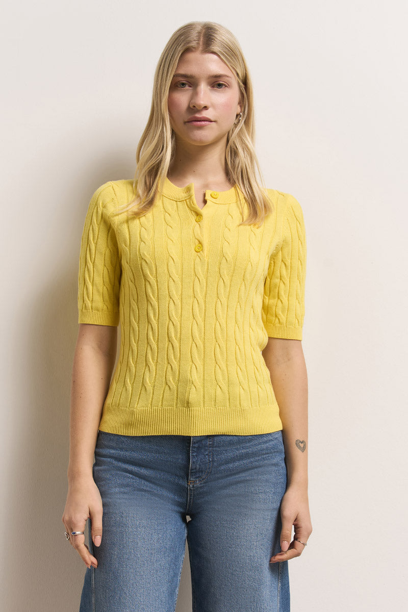 Eine Frau mit langen blonden Haaren trägt einen leuchtend gelben, kurzärmeligen Princess Goes Hollywood Zopfstrick-Pullover aus Baumwolle mit Kragen und Knöpfen, gepaart mit hochgekrempelten Blue Jeans. Sie steht vor einer hellen Wand und blickt neutral nach vorne.