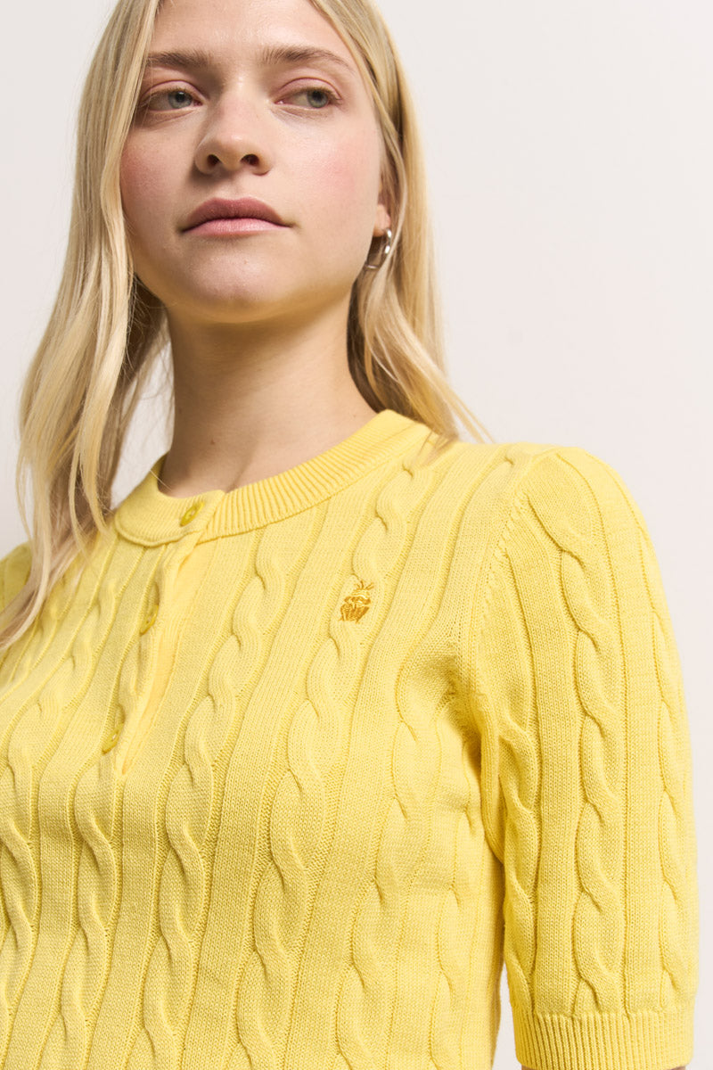 Eine Frau mit langen blonden Haaren trägt einen hellgelben Princess Goes Hollywood Pullover aus Baumwollzopfstrick mit kurzen Ärmeln, Knöpfen und einer kleinen Froschstickerei auf der Brust. Sie blickt leicht zur Seite vor einem schlichten, hellen Hintergrund.