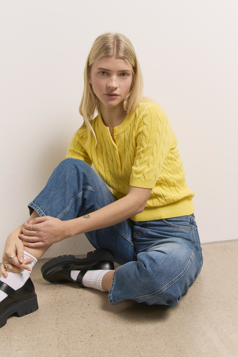 Eine junge Frau mit langen blonden Haaren sitzt auf dem Boden vor einer weißen Wand. Sie trägt einen gelben Baumwollpullover mit Zopfmuster von Princess Goes Hollywood, blaue Jeans, weiße Socken und schwarze, klobige Slipper. Sie blickt entspannt nach vorne, ein Knie hochgezogen und den Arm darauf gestützt.