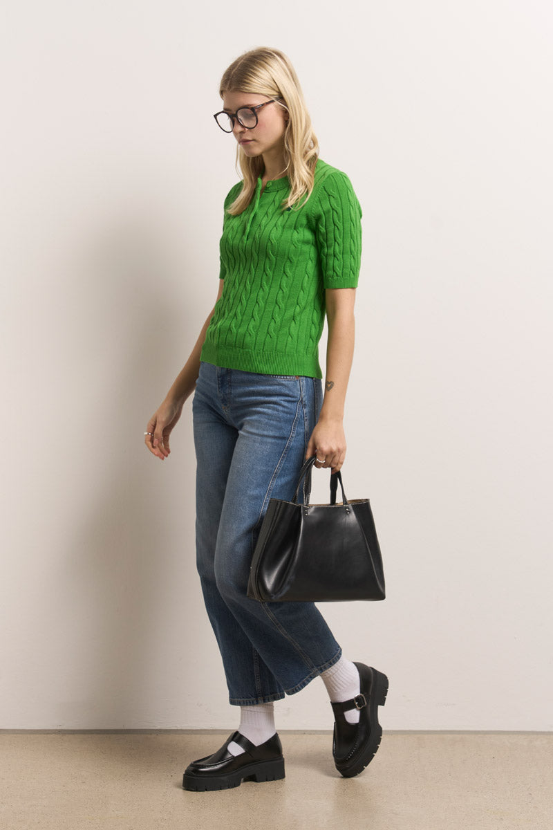 Eine blonde Frau mit Brille trägt einen hellgrünen Princess Goes Hollywood Zopfstrick-Pullover (Baumwolle, kurzärmelig), blaue Jeans mit Bündchen, weiße Socken, schwarze Halbschuhe und eine schwarze Handtasche, während sie vor einer schlichten Wand steht.